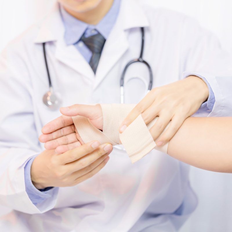 Male doctor putting gauze on young man's hand in clinic, closeup. First aid